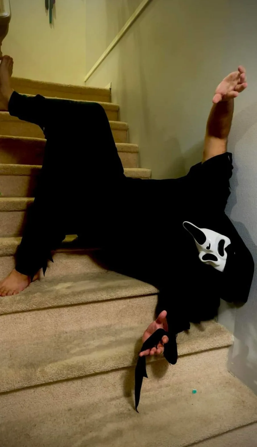 A person wearing a black t-shirt with a white Ghostface mask from the Scream movie franchise is lying on carpeted stairs…