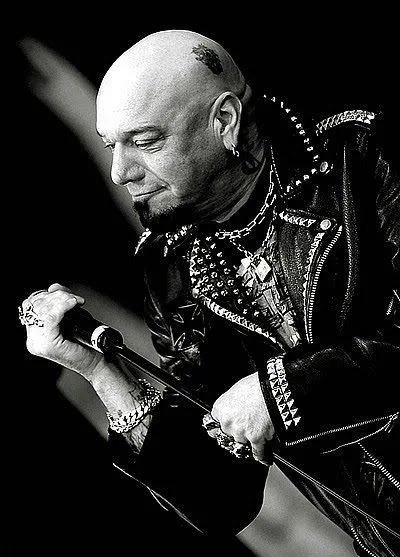 A black-and-white photo of a bald man with a goatee, wearing a heavily decorated leather jacket with studs and chains…