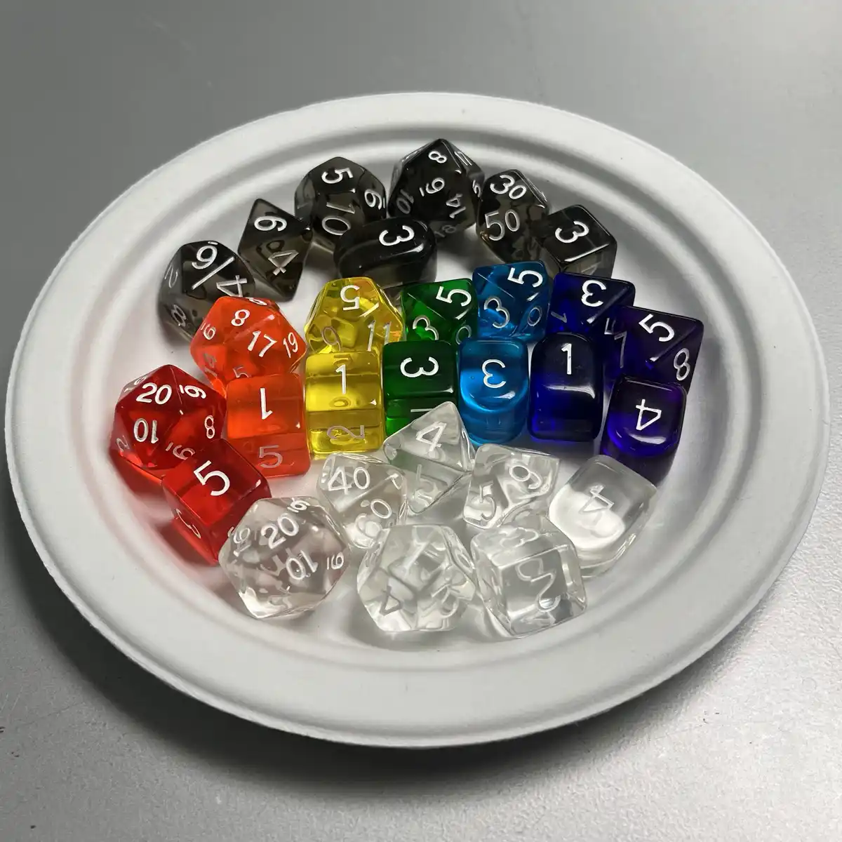 A white paper plate holds a rainbow-colored assortment of polyhedral dice, including red, orange, yellow, green, blue…
