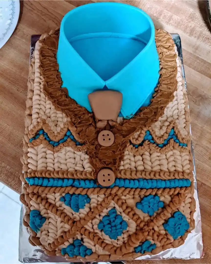 A rectangular cake decorated to resemble a blue-collared shirt with brown and beige frosting piped to look like knitted…