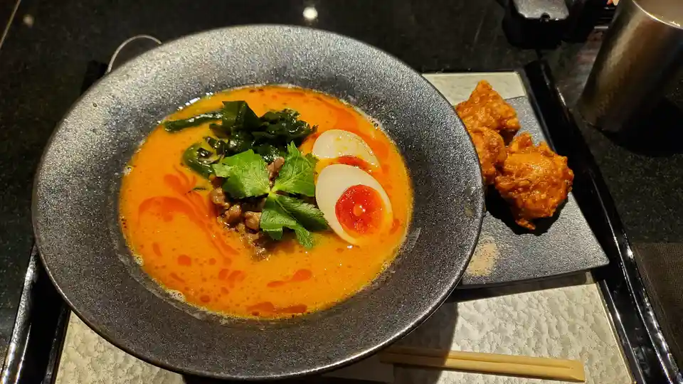 A close-up photo shows a dark gray ceramic bowl filled with ramen featuring an orange broth, a soft-boiled egg sliced in…