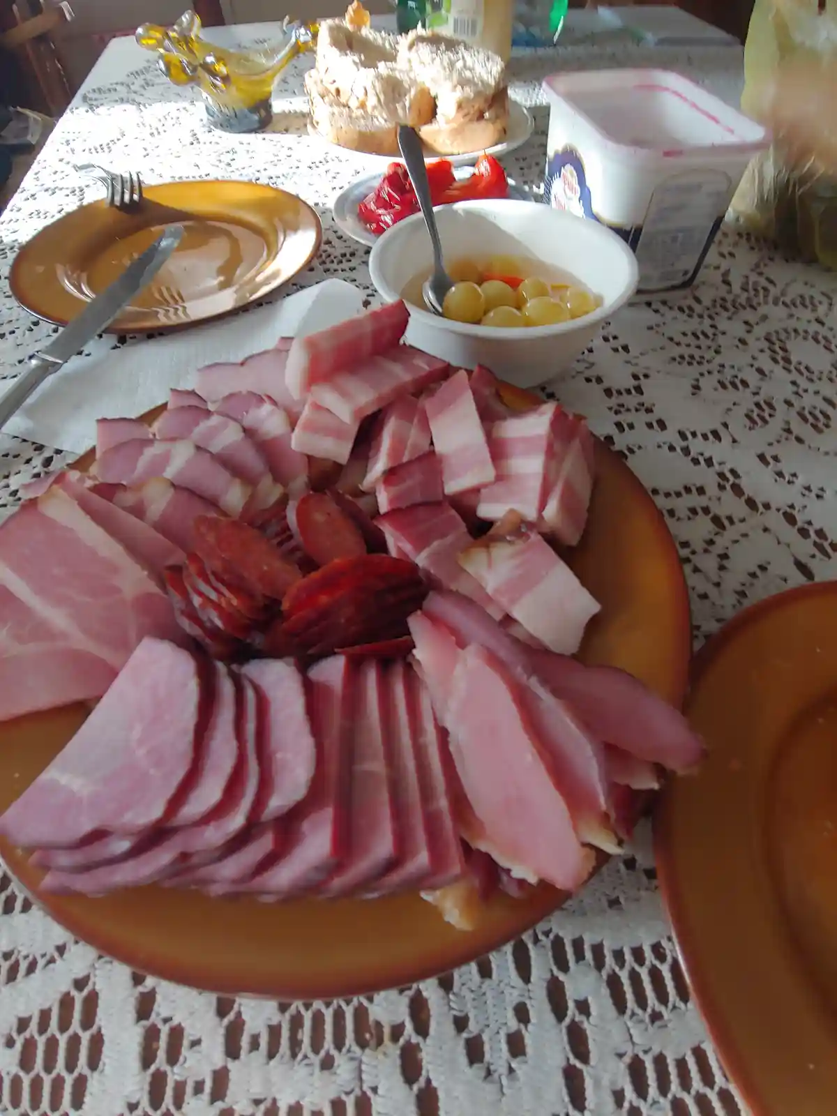 A photo of a large plate of assorted sliced meats including ham, bacon, and salami, arranged on a lace tablecloth with other…