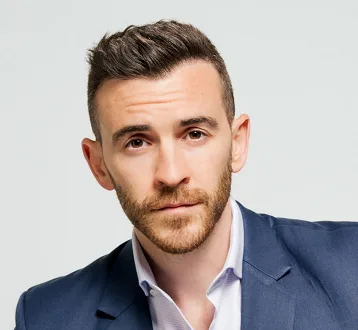 A man with short dark hair and a beard, wearing a blue suit jacket over a light blue collared shirt, stares directly at the…