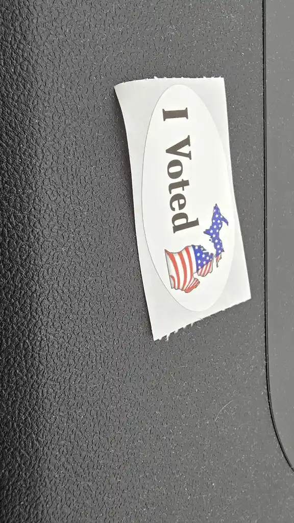 A close-up of a white oval 'I Voted' sticker with a patriotic design featuring the outline of the state of Massachusetts…
