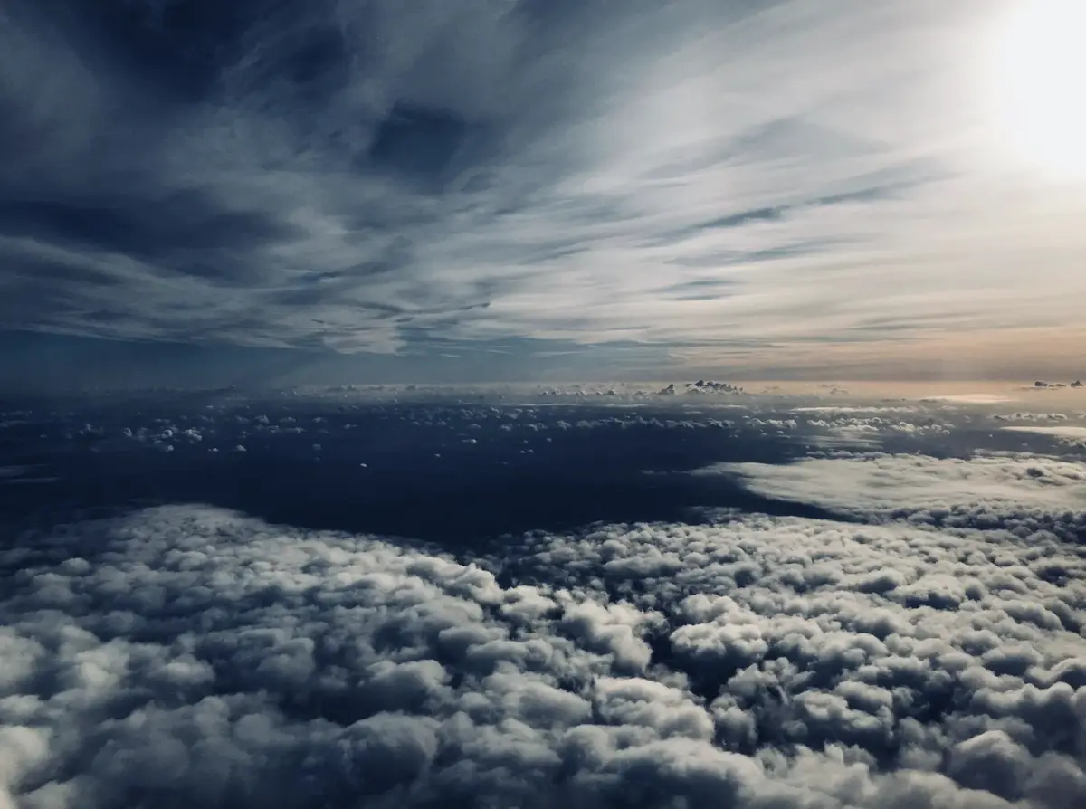 An aerial view from above a dense layer of fluffy white clouds, with darker storm clouds and a bright sun visible in the…