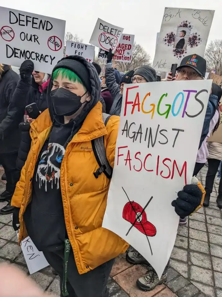 A person with green hair and a black face mask holds a sign reading 'FAGGOTS AGAINST FASCISM' with a crossed-out red MAGA…
