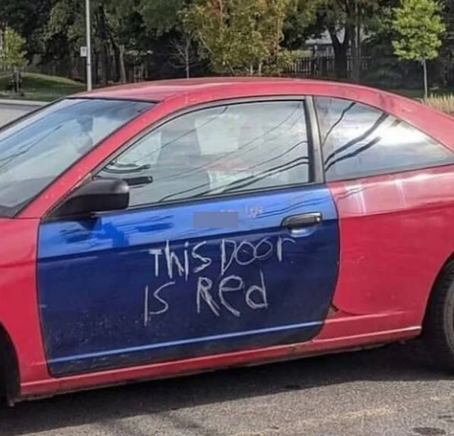 A red car has its driver's side door painted blue, with the phrase 'THIS DOOR IS RED' written in white chalk on the blue…
