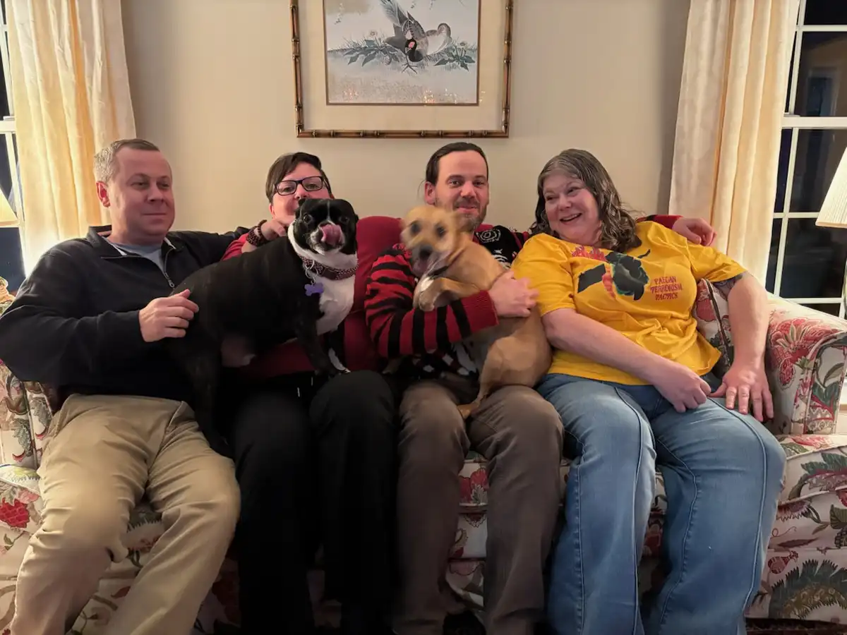 four people are sitting on a floral-patterned couch in a living room, each holding a dog. The person on the far left holds a…