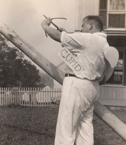 A black-and-white photo shows a man in a light-colored shirt and pants, standing outdoors and aiming a bow and arrow. He…