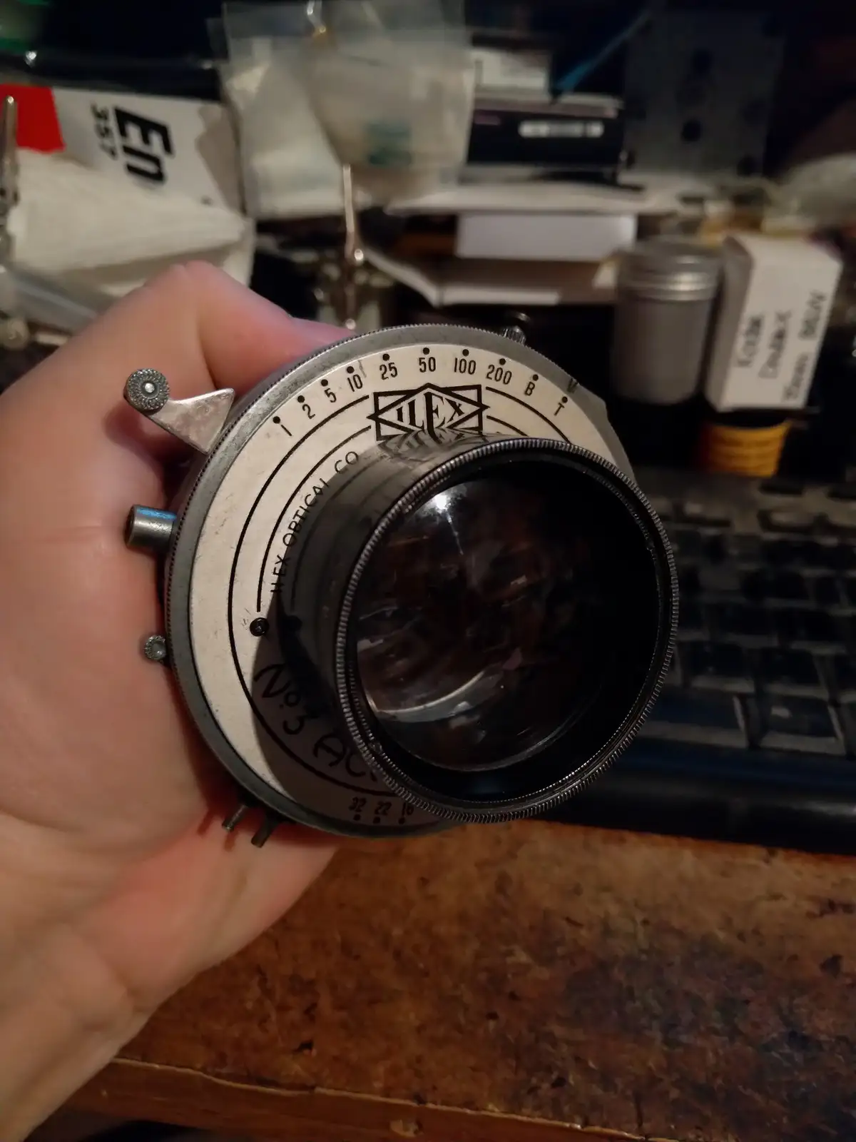 A hand holds an old Ilex Optical Co. No.3 Auto camera lens, showing its engraved markings and aperture settings, against a…