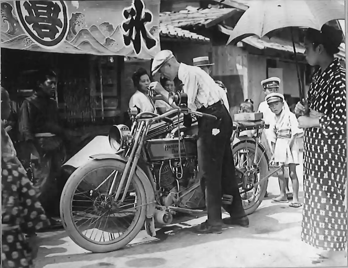 A black-and-white historical photo shows a man in a cap leaning over a vintage Harley-Davidson motorcycle, adjusting its…