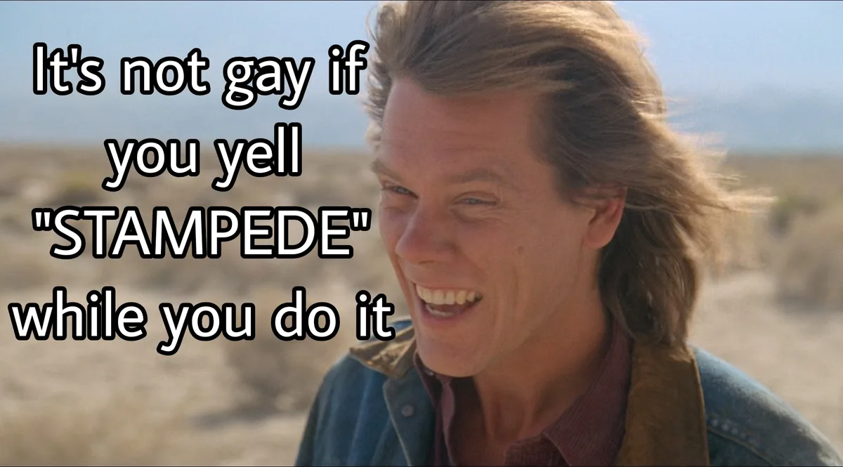 A man with long brown hair, wearing a denim jacket over a collared shirt, is shown laughing broadly outdoors in a…