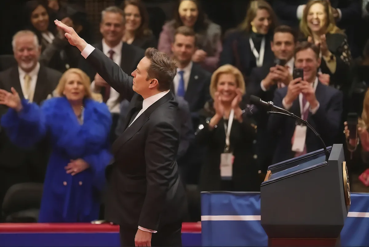 A man in a black suit stands at a podium with his right arm raised, facing an audience of smiling people who are applauding…