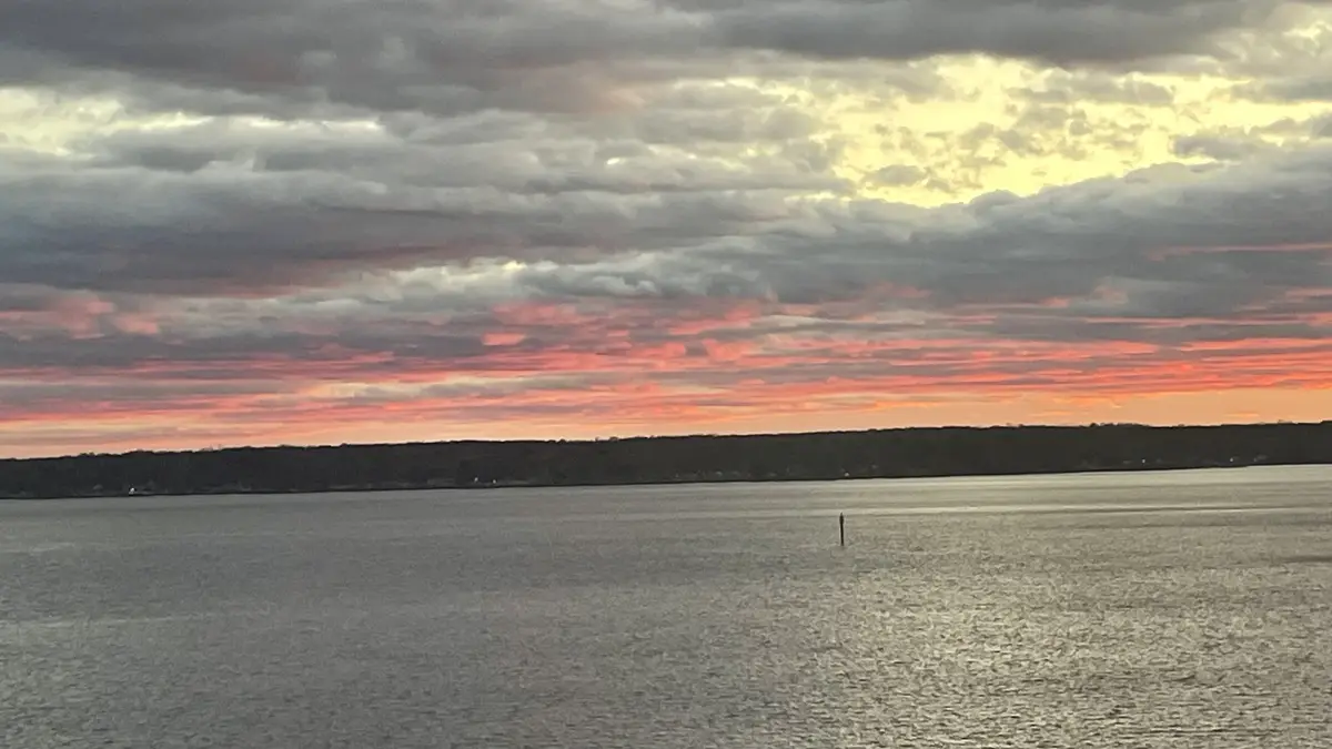 A serene sunset over a large body of water with a distant shoreline, featuring dramatic clouds painted in shades of orange…
