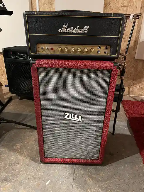 A black Marshall guitar amplifier head sits atop a speaker cabinet with a red, textured, crocodile-skin-like exterior. The…
