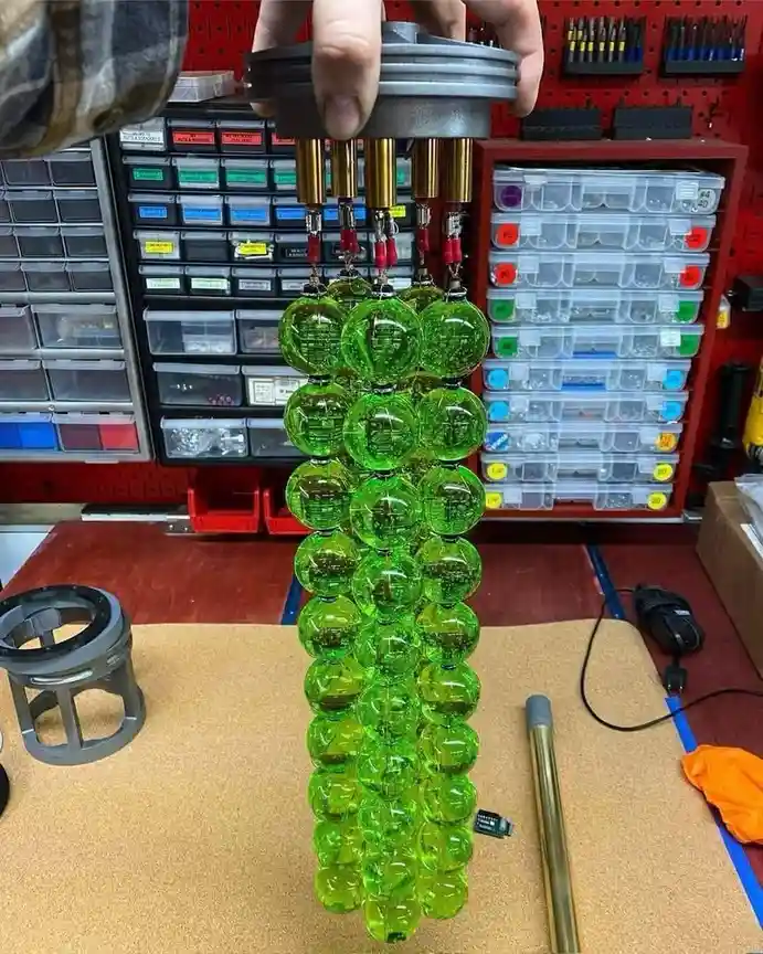 A person holds a gray mechanical top piece above a vertical tower of 30+ translucent green spheres, each containing visible…
