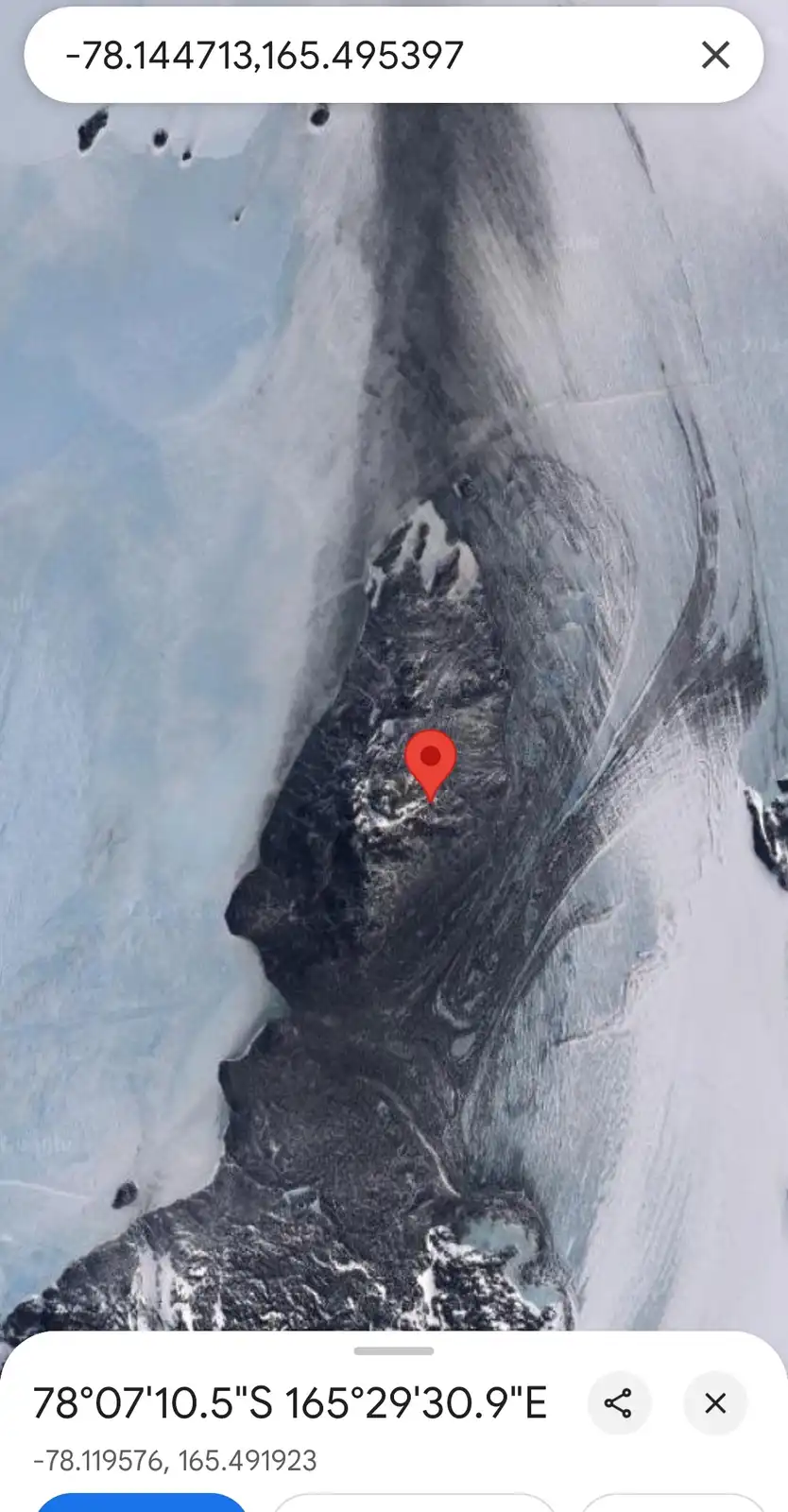 A satellite map view shows a rugged, snow-dusted mountain range surrounded by ice and glaciers, with a red location pin…