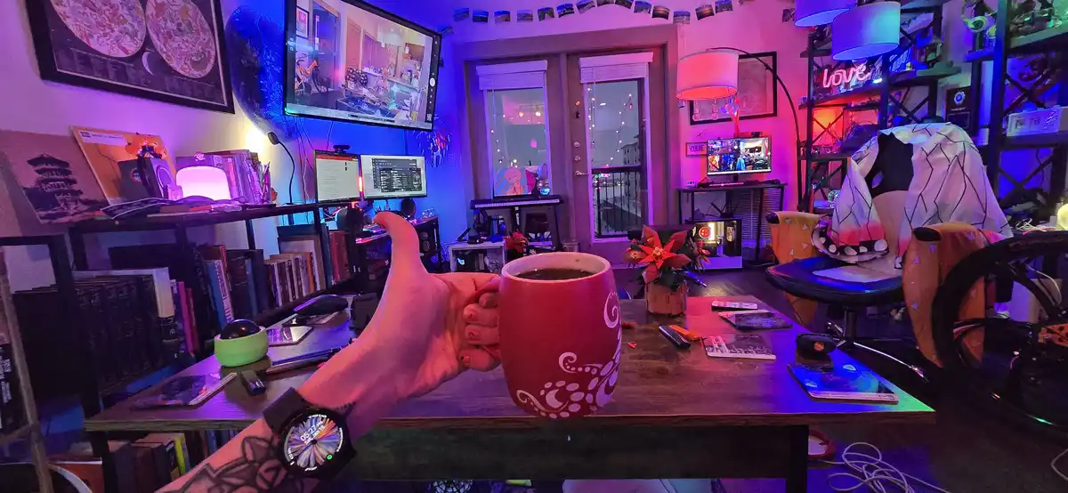 A hand holds a red mug with white swirls, giving a thumbs-up gesture in front of a cluttered, neon-lit streaming room…