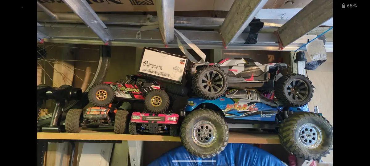A cluttered shelf in a garage holds several remote-controlled monster trucks and a box labeled '43 CHASSIS MODEL 1:43 RC…