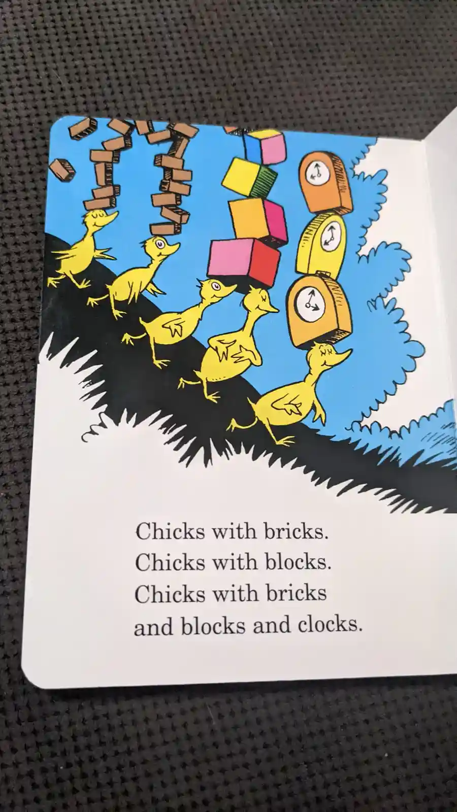 A children's book illustration shows five yellow chicks running along a black hillside, each balancing a stack of items on…