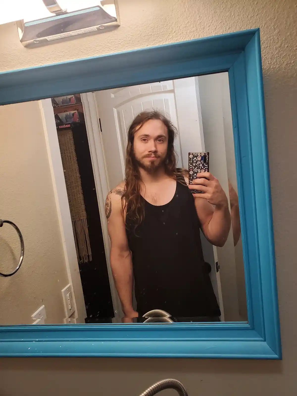 A man with long hair and a beard takes a mirror selfie in a bathroom, wearing a black tank top and holding a phone with a…