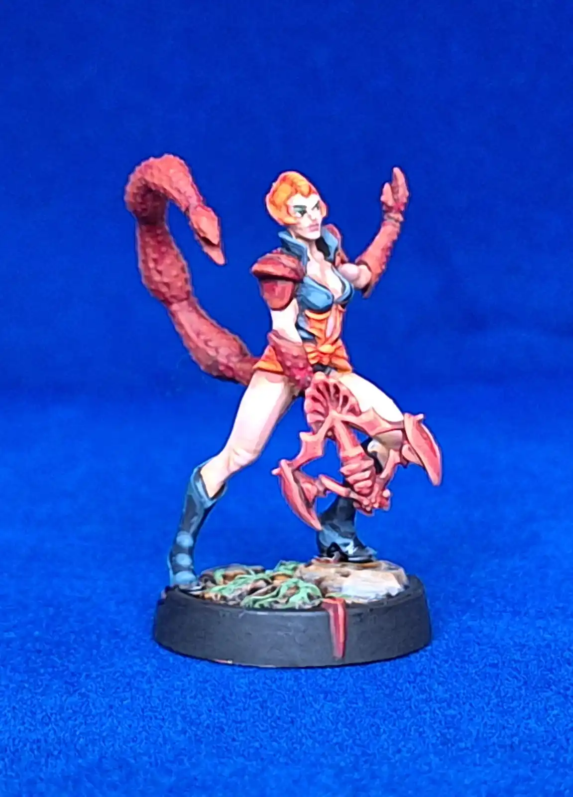 A painted miniature figurine of a female scorpion warrior with orange hair, red scorpion tail, and scorpion-like armor…