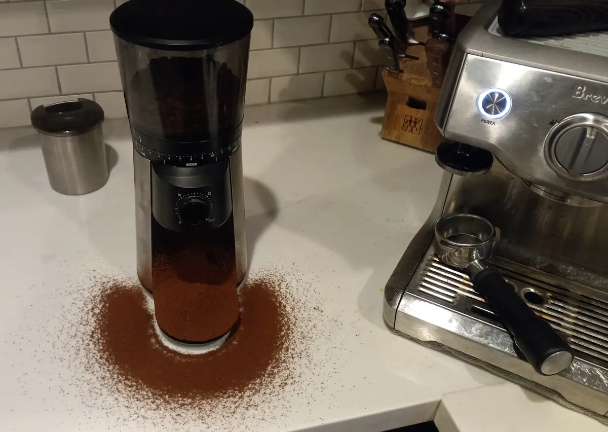 A coffee grinder sits on a white kitchen counter with a large spill of ground coffee around its base, next to a stainless…