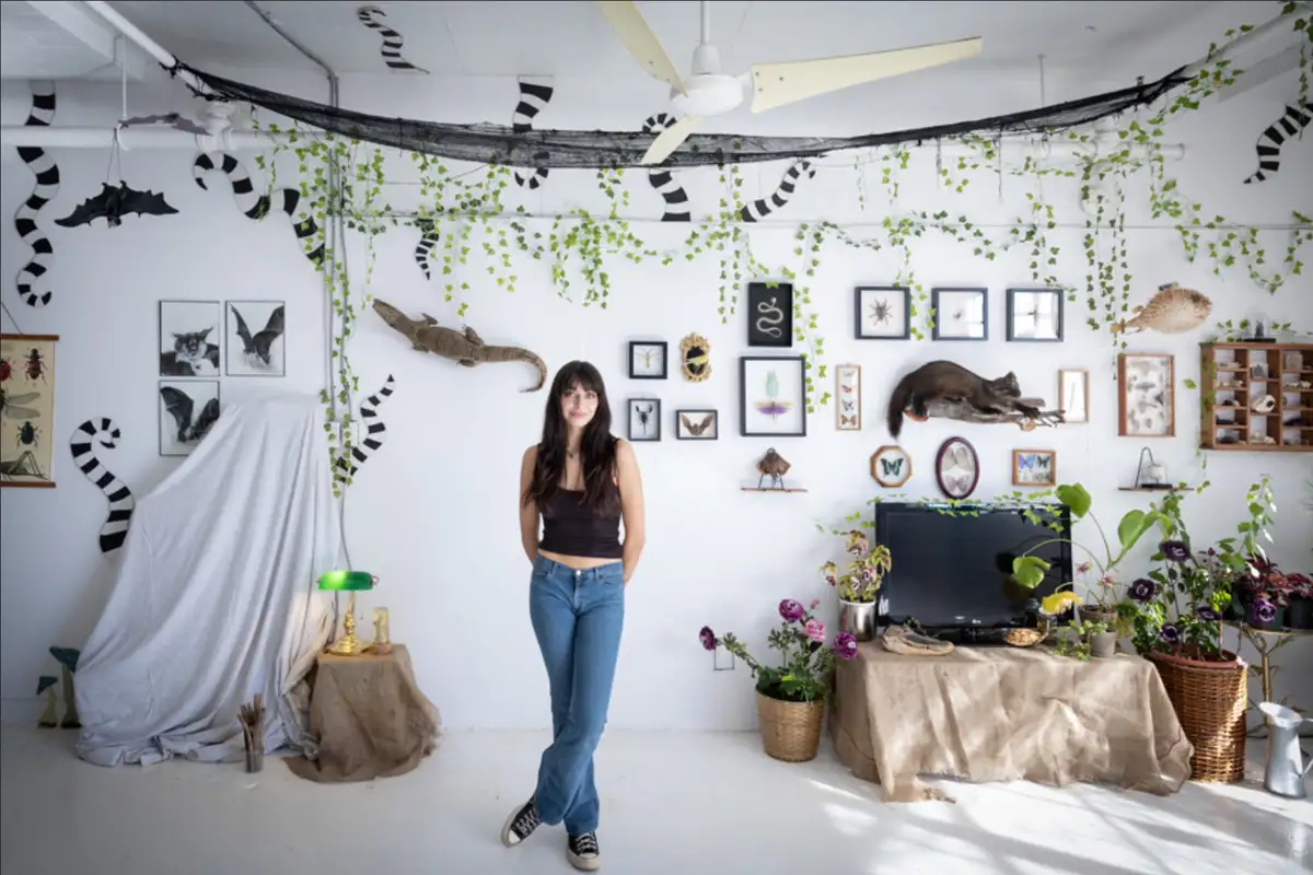A woman stands in a room decorated with numerous taxidermy animals including bats, a crocodile, and a lemur, along with…