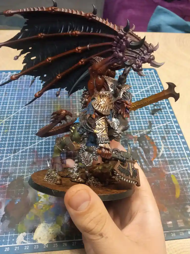A hand holds a highly detailed, painted fantasy miniature of a muscular, horned, winged creature resembling a dragon or…
