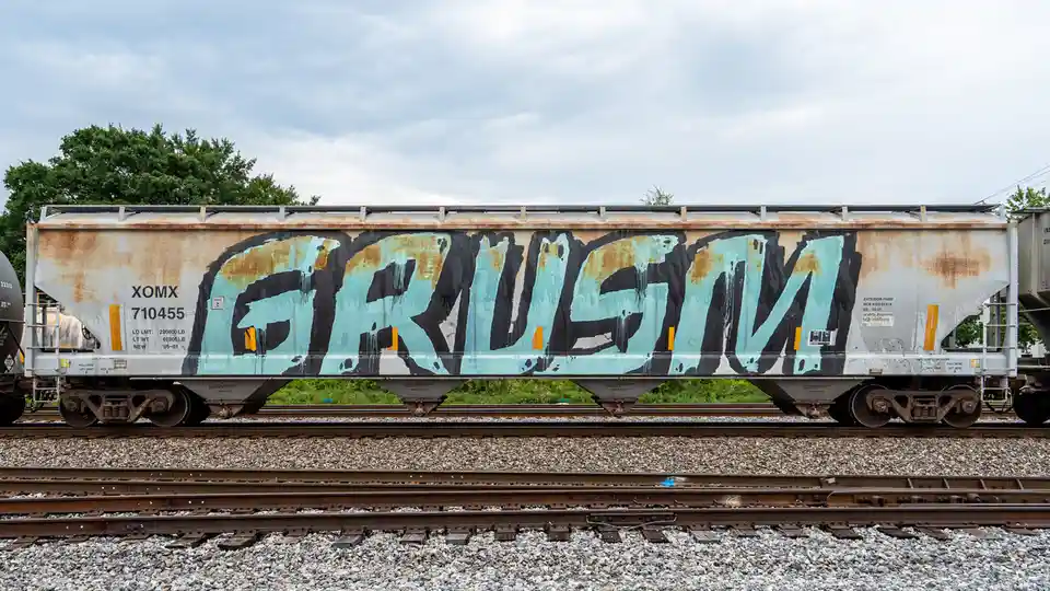 A weathered silver railroad hopper car sits on tracks under a cloudy sky, featuring large, stylized graffiti that spells…