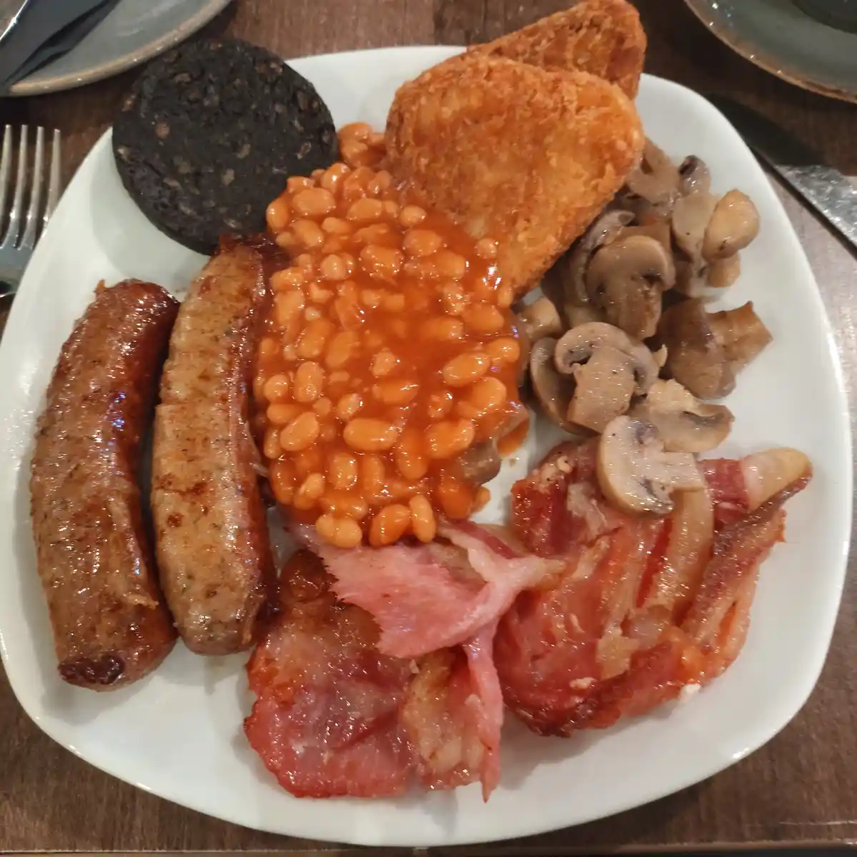 A plate of traditional full english breakfast featuring two sausages, bacon, baked beans, fried mushrooms, hash browns, and…