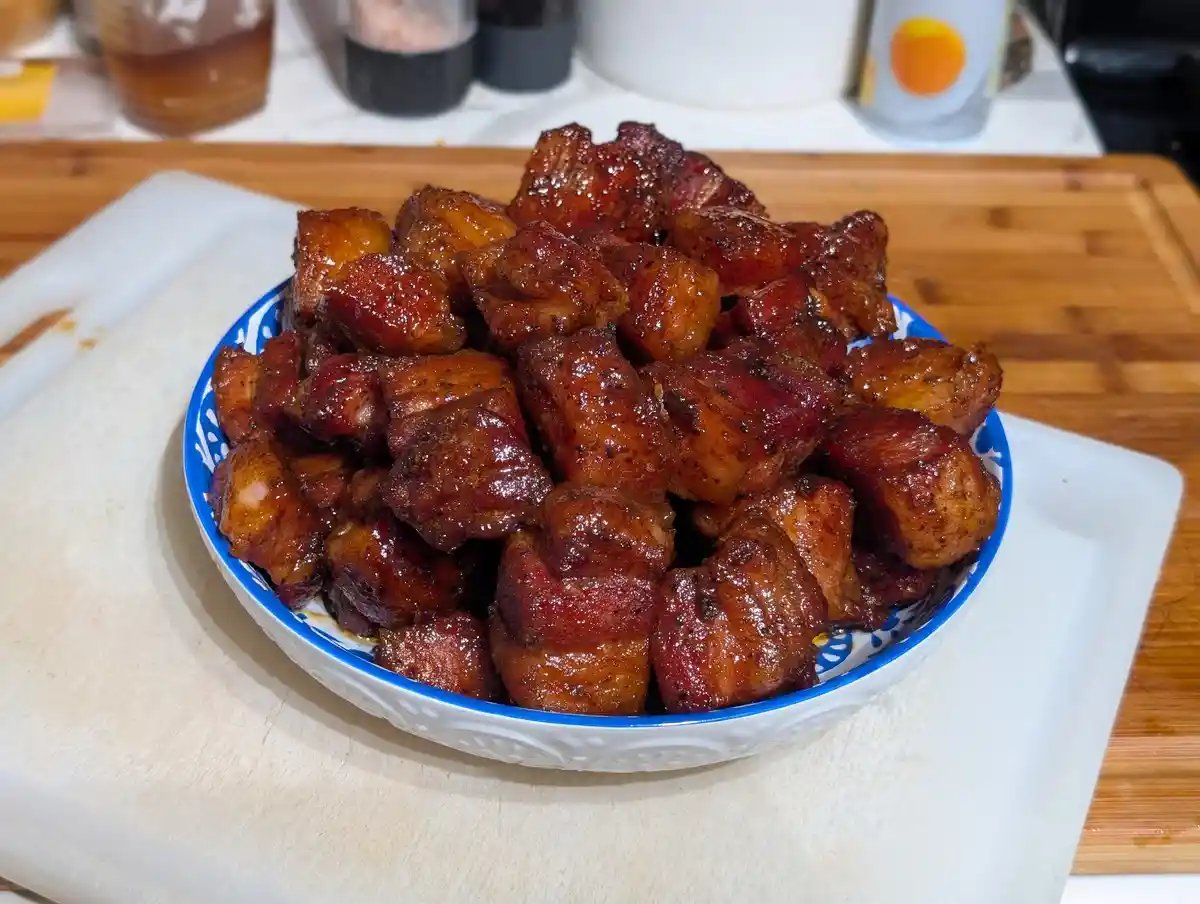 A white ceramic bowl with a blue rim and patterned interior is filled with glossy, caramelized cubes of cooked meat, likely…