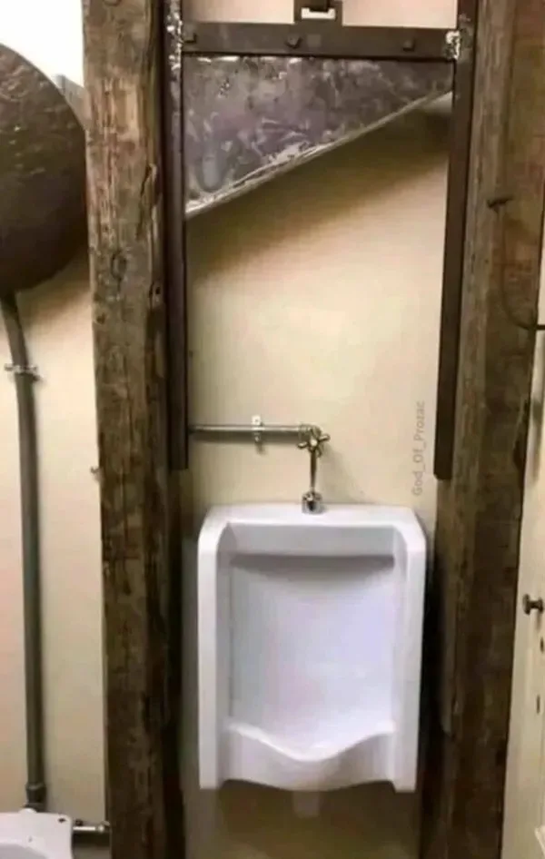 A white urinal is mounted on a beige wall between two weathered wooden beams in a rustic bathroom. Above the urinal, a metal…
