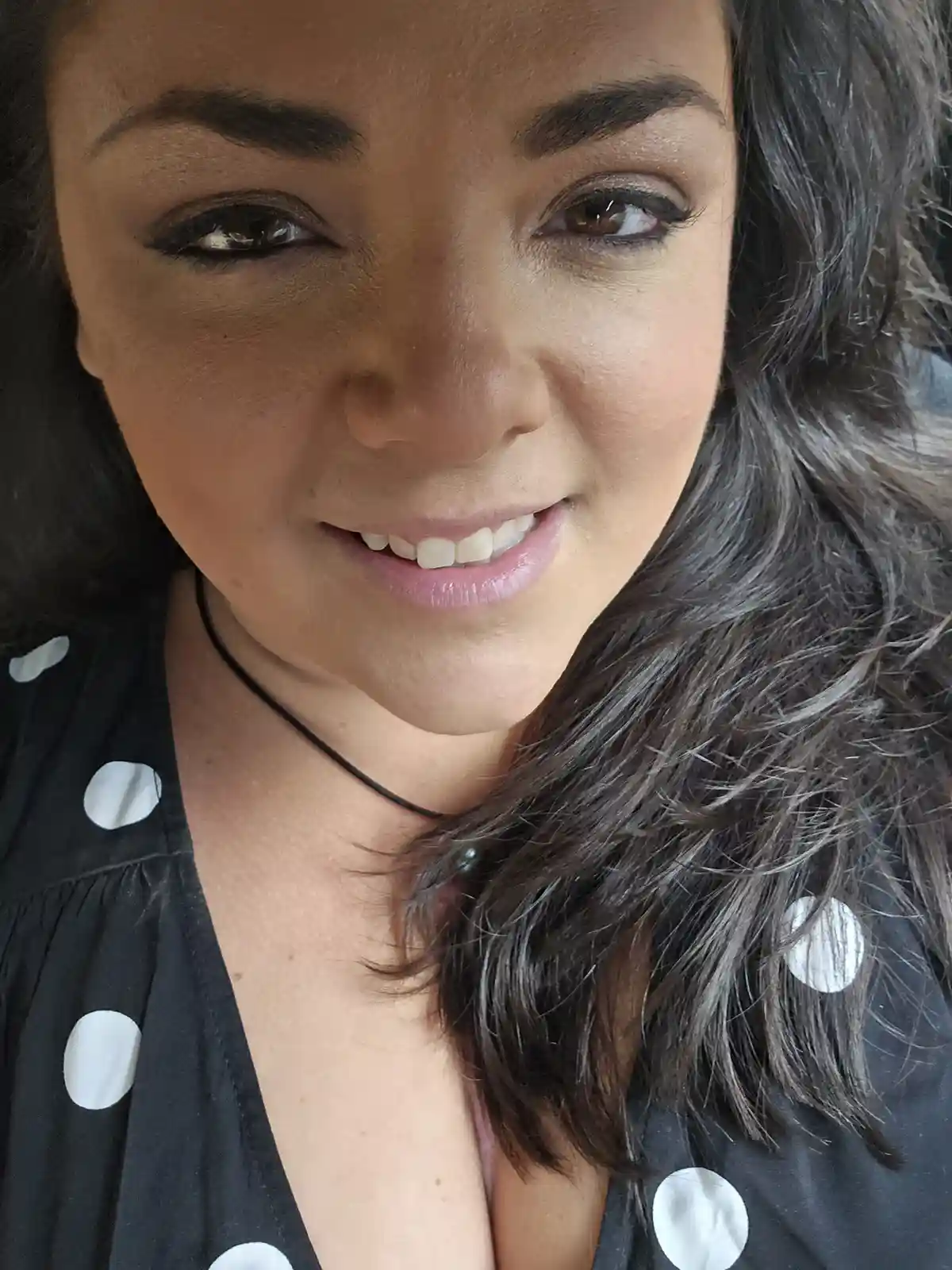 A close-up selfie of a woman with dark, wavy hair and makeup, smiling slightly while wearing a black top with white polka…