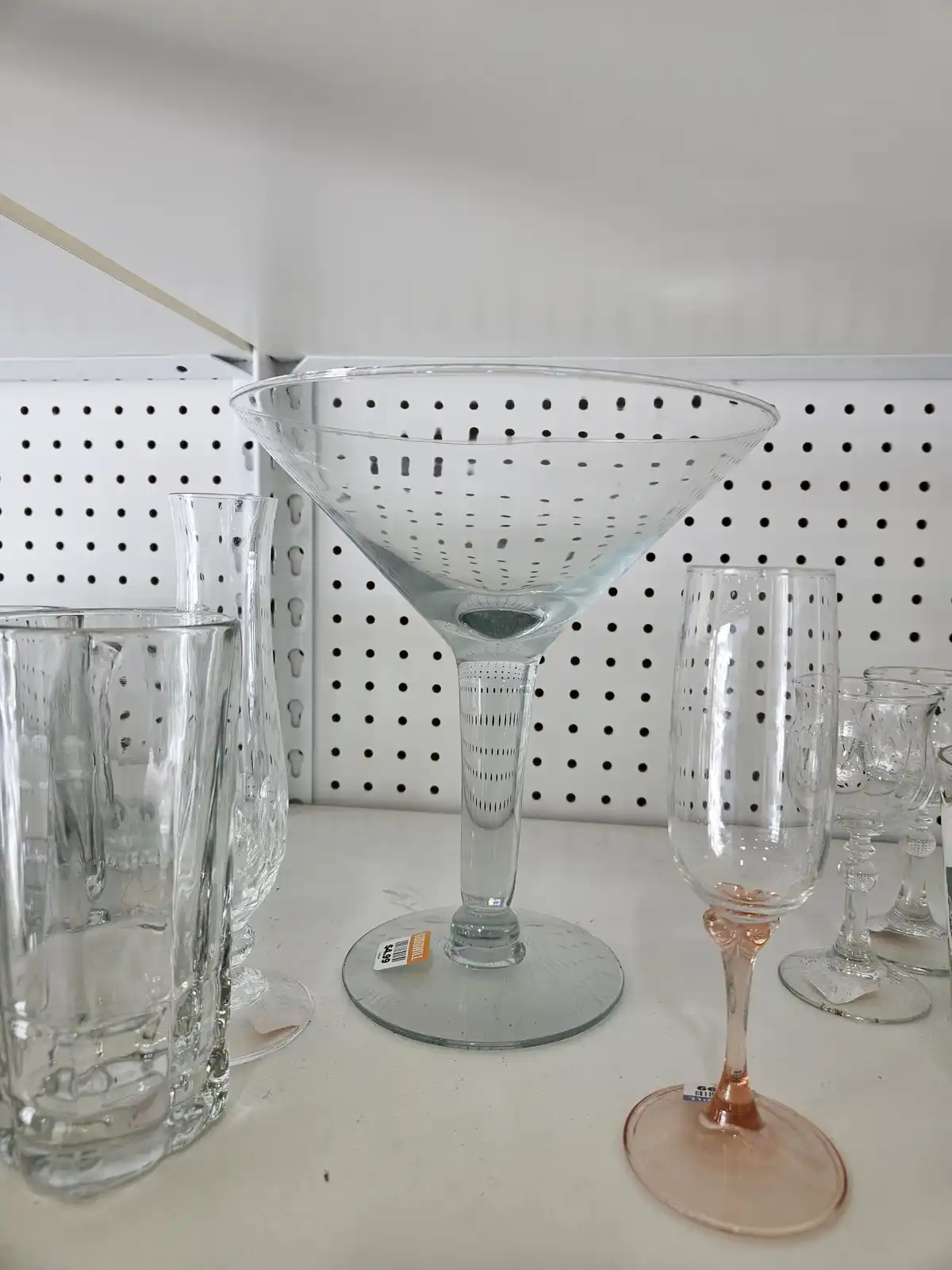 a martini glass with holes punched through its bowl sits on a store shelf among other glassware, with a price tag visible…