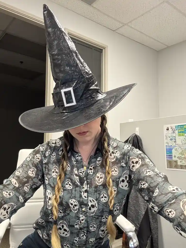 A person wearing a large, shiny black witch's hat that obscures their face, seated in an office chair. They are wearing a…
