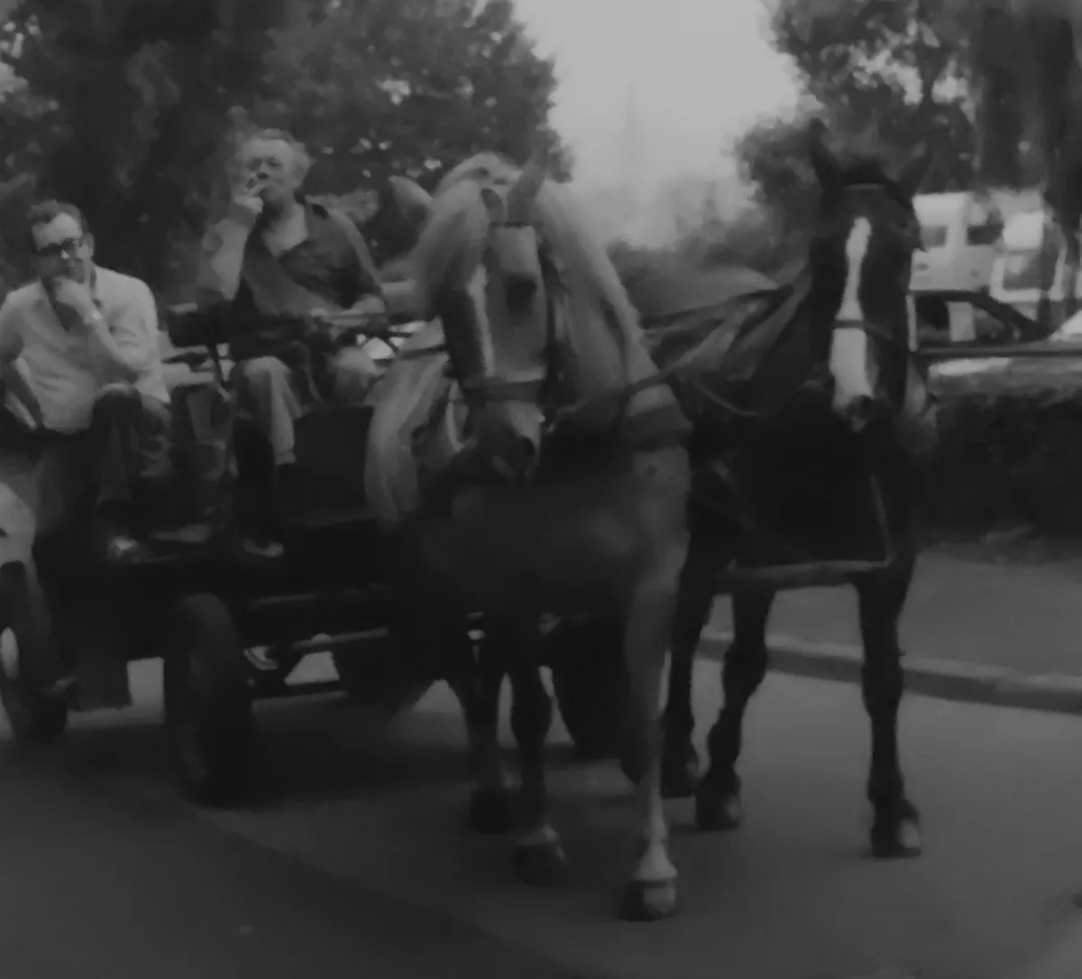 A blurry black-and-white photo shows two men sitting in a horse-drawn carriage with two horses. One man is smoking a…