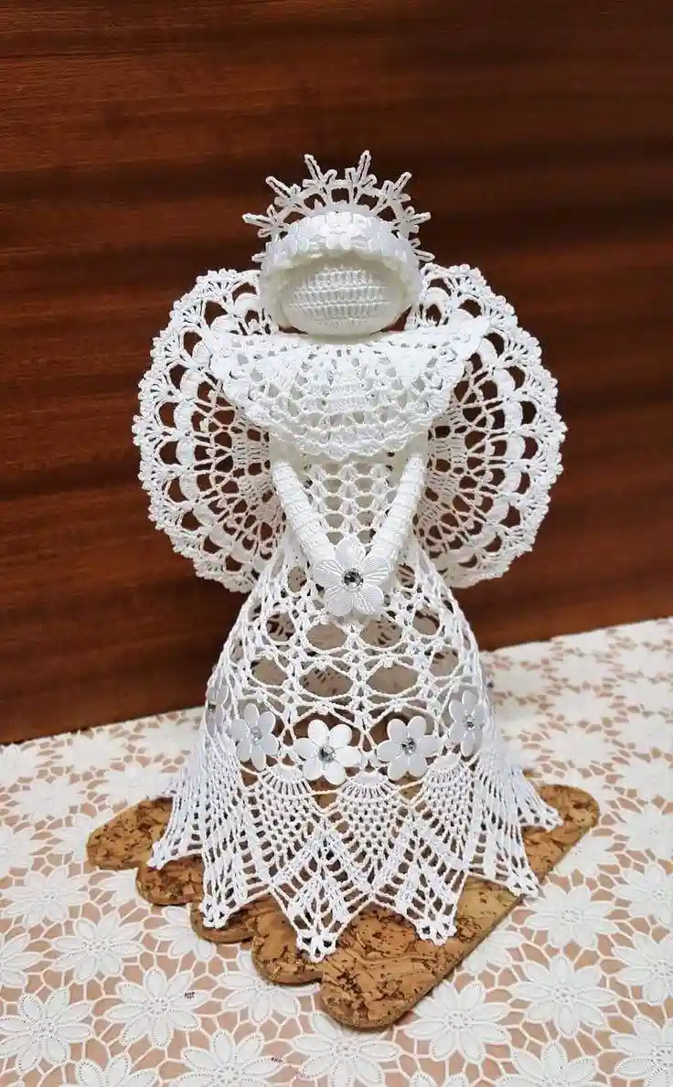 A white crocheted angel figurine with intricate lace wings and dress, but its feet are made of cork coasters, placed on a…