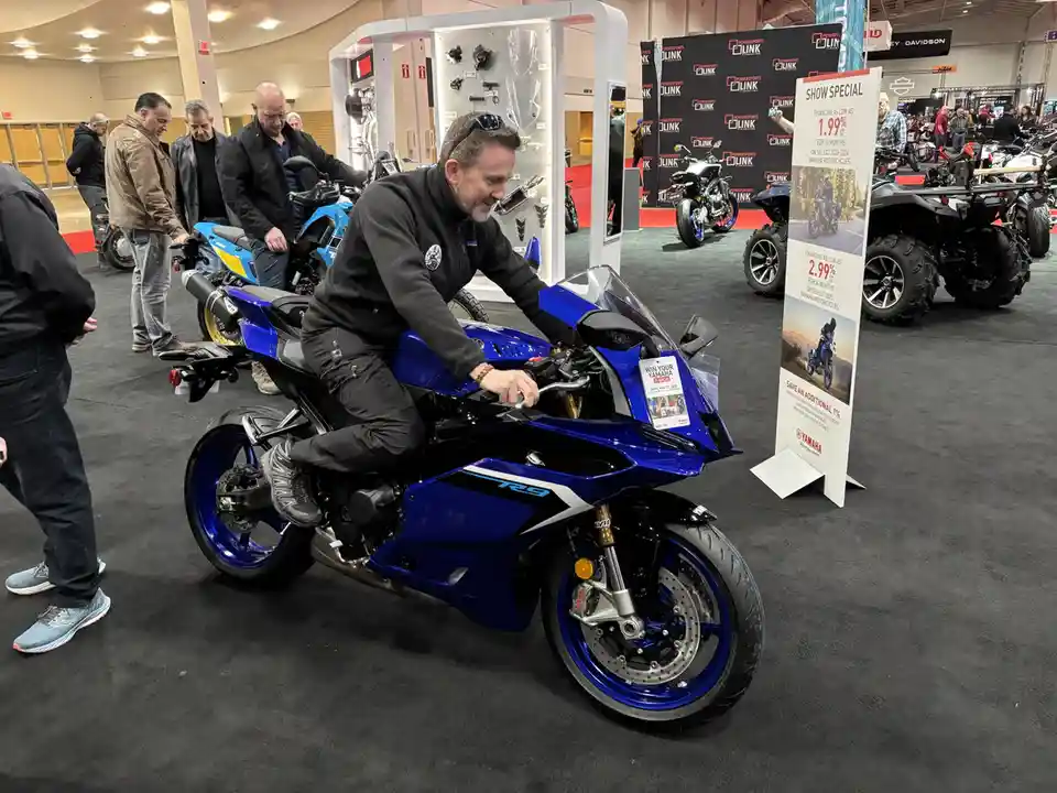 A man in a black jacket sits on a blue Yamaha R9 sport motorcycle at a trade show, smiling as he touches the handlebars…