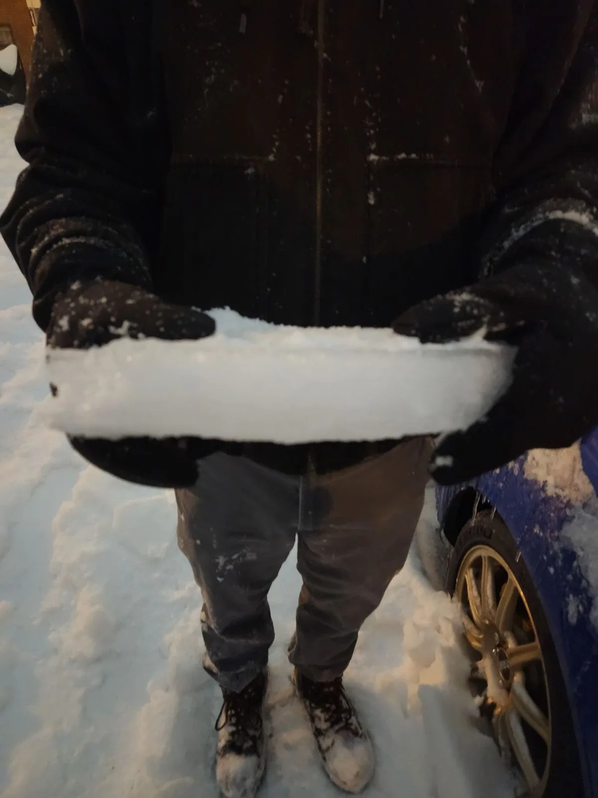 A person wearing a dark jacket, black gloves, and boots stands in snow, holding a thick, rectangular block of ice like a…