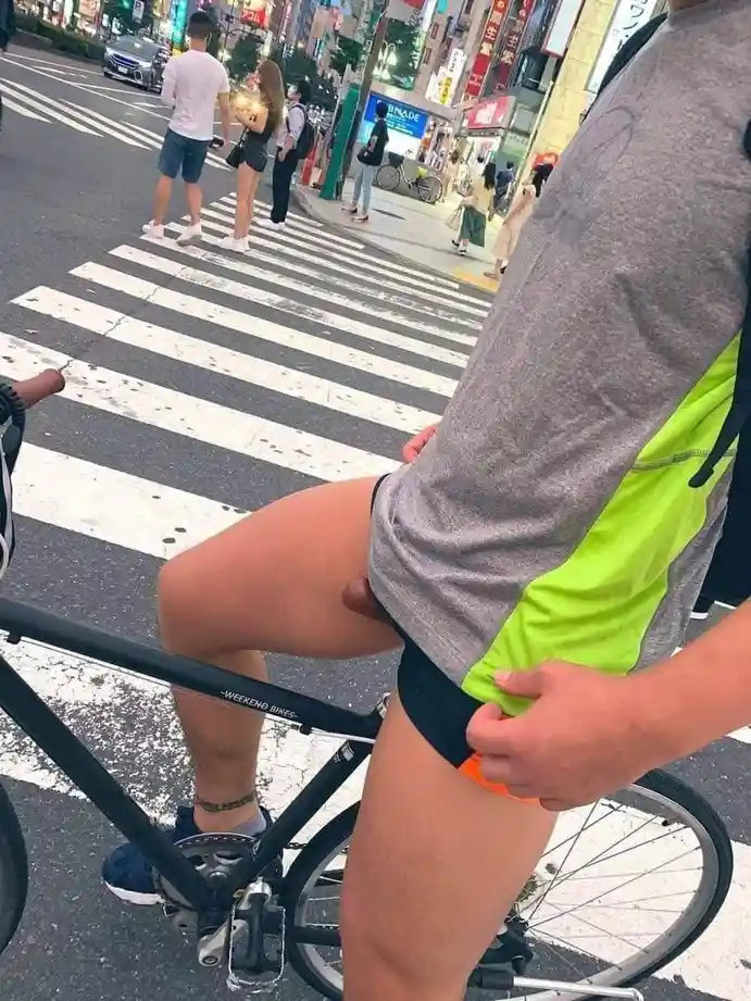 A man on a black bicycle at a city crosswalk is adjusting his black underwear, revealing orange briefs underneath, while…