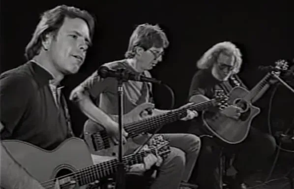 A black-and-white photo of three men playing acoustic guitars on stage, with the man on the left singing into a microphone…