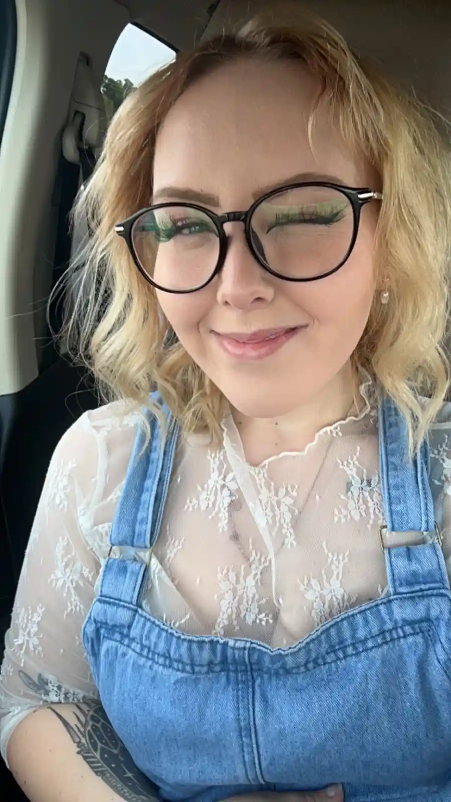 A woman with blonde hair and black-rimmed glasses is sitting in a car, smiling slightly with one eye closed. She wears blue…