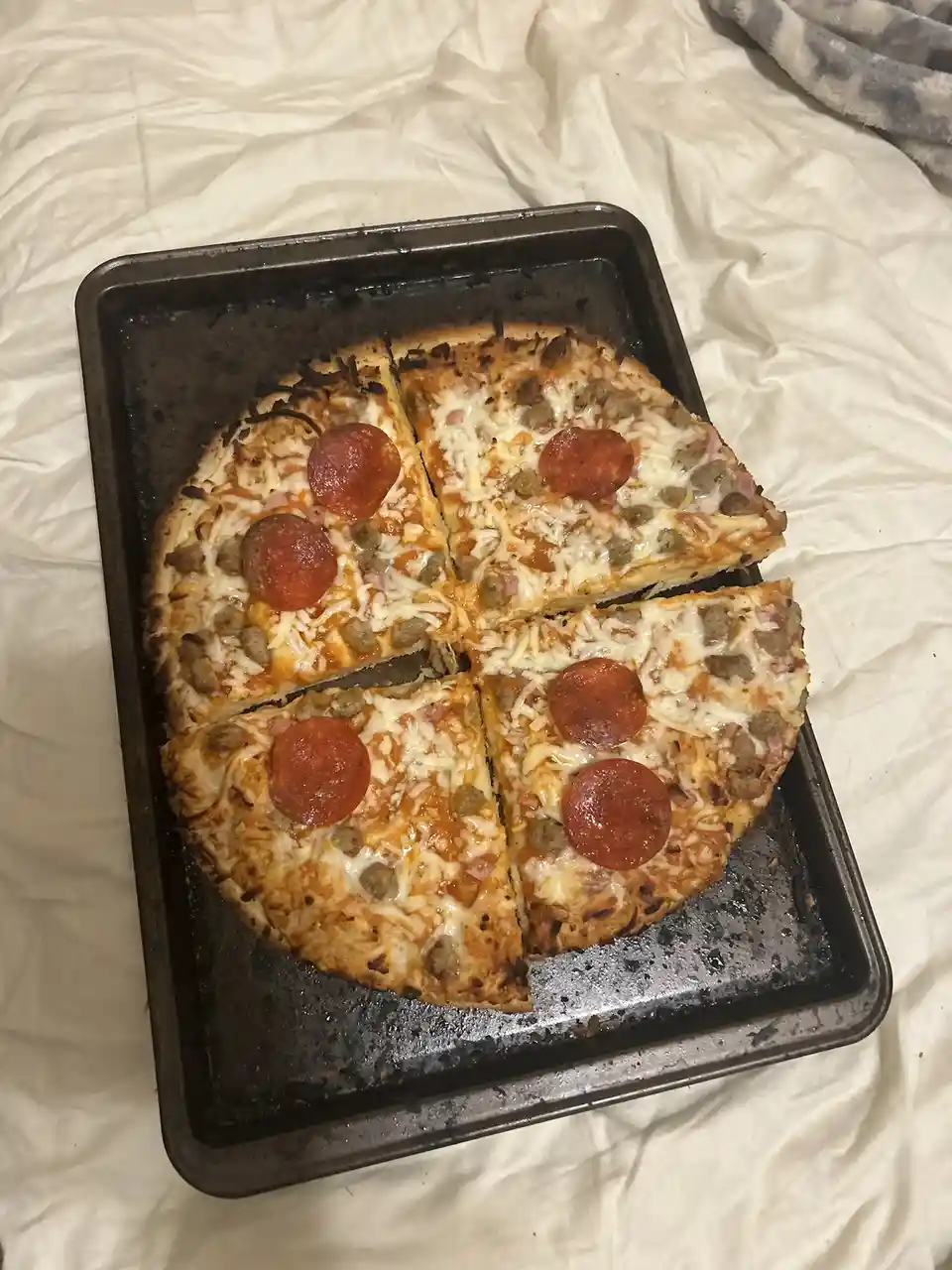 A homemade pizza with pepperoni and sausage toppings, sliced into four pieces and resting on a dark baking sheet, which is…