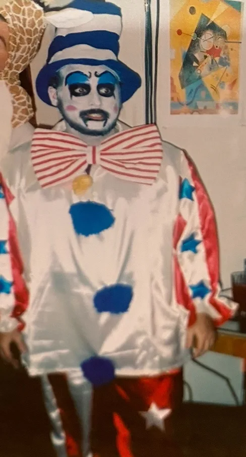 A person in a full clown costume with a blue and white striped hat, heavy blue eye makeup, a large red and white striped bow…