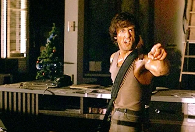 a man with curly hair and a tank top, holding a rifle over his shoulder, points his finger forward with an intense…