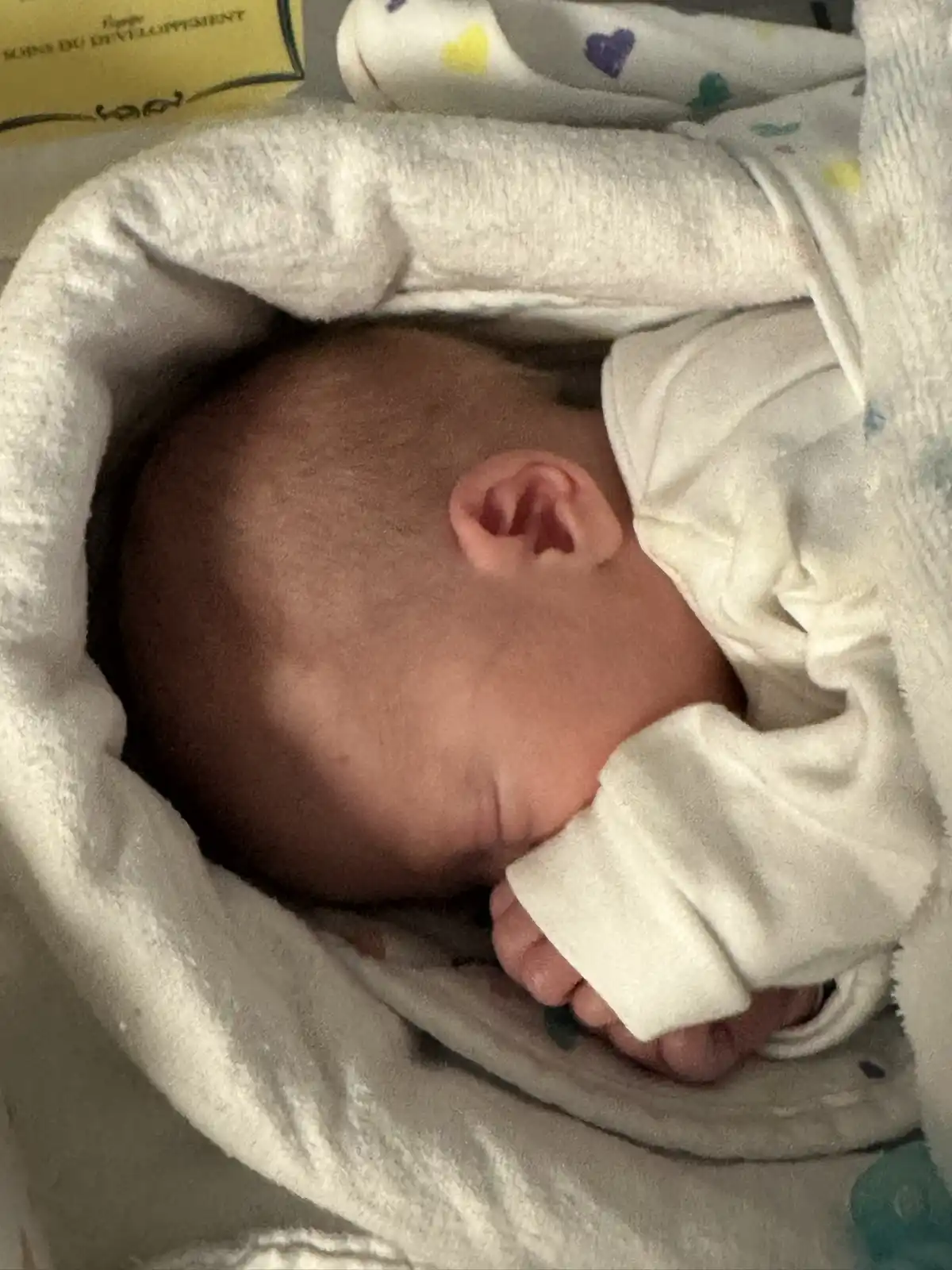 A close-up photo shows a newborn baby's head and one ear peeking out from a white, fluffy blanket. The baby is wearing a…