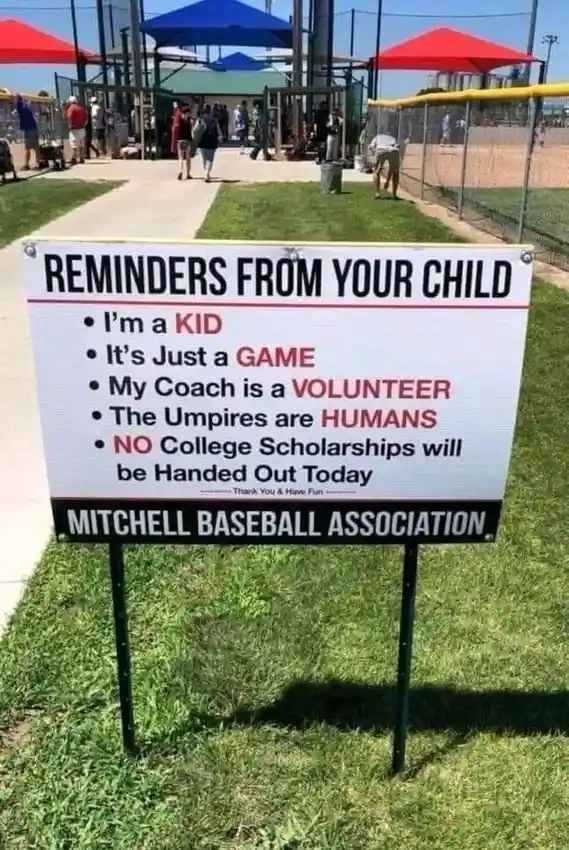 A white sign with black and red text stands on grass at a baseball field, listing humorous reminders from a child to…