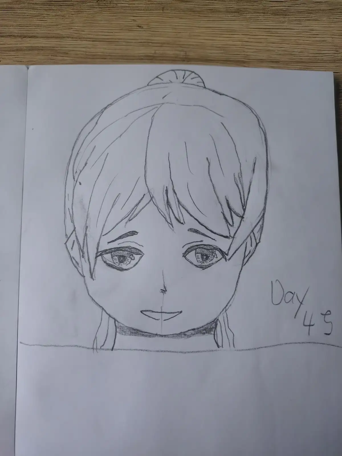 A hand-drawn pencil sketch on paper shows a stylized anime-style face with short hair and large eyes, resting on a wooden…