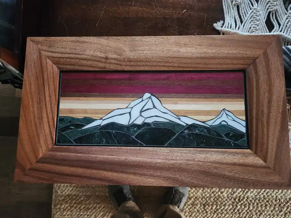 A wooden-framed piece of art depicting a stylized mountain range with snow-capped peaks, set against horizontal stripes of…